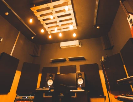 Estúdio de música com teto e paredes marrons, luminária de pallets, monitores de áudio e absorvedores de som. Ambiente iluminado por lâmpadas pendentes.