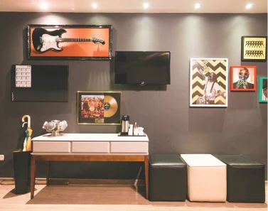 Sala com decoração musical inclui guitarra na parede, TV, disco de ouro emoldurado e pôster. Mesa branca, sofá e iluminação aconchegante.