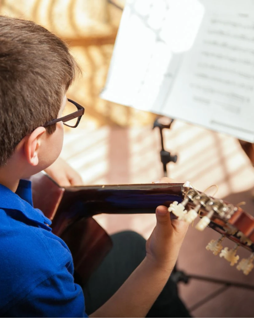 Garoto com óculos lê partitura e toca violão clássico. Camisa azul e luz natural suave.
